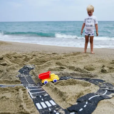 Petit garçon sur la plage, jouant avec un circuit routier en bandes noires et blanches dans le sable, voiture jaune et rouge en place près de l’océan. Circuit de route flexible Waytoplay, coffret Autoroute 24 pcs.