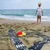 Petit garçon sur la plage, jouant avec un circuit routier en bandes noires et blanches dans le sable, voiture jaune et rouge en place près de l’océan. Circuit de route flexible Waytoplay, coffret Autoroute 24 pcs.
