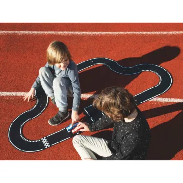 Deux enfants jouent avec un circuit de course noir sur une piste rouge, assemblé comme un parcours sinueux.