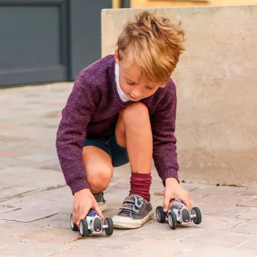 Jeune garçon en chemise manches longues violette et short, prêt à lancer deux petites voitures de course sur le trottoir.