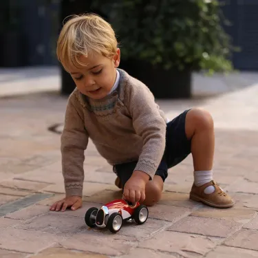 Petit garçon jouant avec une voiture miniature rouge et blanche sur un sol en pavés.