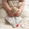 Doudou lapin SIGIKID 31269 en tissu éponge beige et coton fleuri avec oreilles longues, tenu par des mains d’enfant sur une couverture tricot beige