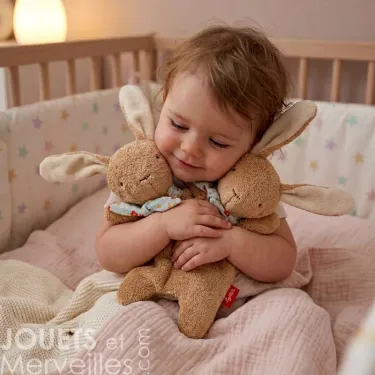 Jolie bébé dans un lit, câlinant deux peluches lapins marron SIGIKID 31268 en tissu éponge beige, avec couverture rose.