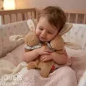 Jolie bébé dans un lit, câlinant deux peluches lapins marron SIGIKID 31268 en tissu éponge beige, avec couverture rose.