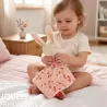 Doudou lapin SIGIKID 31274 en tissu rose à fleurs, bébé assis sur le lit tient la peluche avec son drap floral.
