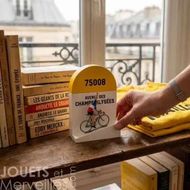 Borne kilométrique Bernard et Eddy - Champs-Élysées, décor de bureau, imprimée "75008 AVENUE DES CHAMPS-ÉLYSÉES" avec figurine de cycliste, posée sur une étagère à côté de livres et d’un maillot jaune.