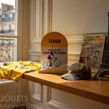 Borne kilométrique jaune et blanche 75008 avec figurine de cycliste et inscription Avenue des Champs-Élysées, affiches et casquette sur une table près d’une fenêtre.