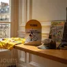 Borne kilométrique jaune et blanche 75008 avec figurine de cycliste et inscription Avenue des Champs-Élysées, affiches et casquette sur une table près d’une fenêtre.