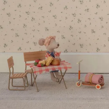 Un souris Maileg est assise sur un banc en bois devant une table de pique nique avec nappe à carreaux et quelques aliments posés sur la table.