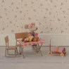 Un souris Maileg est assise sur un banc en bois devant une table de pique nique avec nappe à carreaux et quelques aliments posés sur la table.