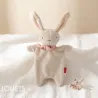 Doudou lapin blanc SIGIKID 31273 en tissu éponge avec collier rose à motifs, debout sur drap blanc.