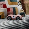 Voiture de course Viglietta Playforever Dottie en rose vintage sur une housse à rayures, numéro 9, et coussins colorés en arrière-plan.