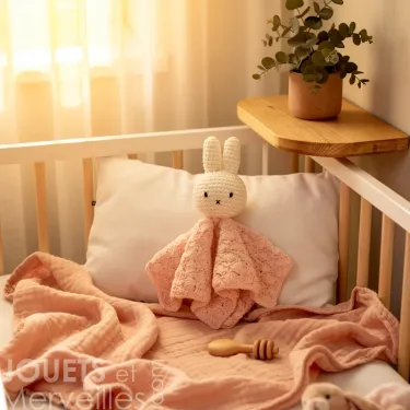Doudou Miffy en crochet - Just Dutch - rose pastel: un chevalier gris? Non, un lapin crocheté blanc assis dans un lit à barreaux, entouré d’une couverture rose pastel et d’un tissu assorti. Pompon et picot lumineux. Plante en pot sur une étagère en bois à côté.