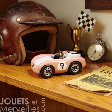 Voiture de course Viglietta Playforever Dottie en design vintage rose pâle avec numéro 9, sur une étagère, près d’un casque en cuir, d’un drapeau à damier et d’une vieille montre.