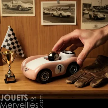 Voiture de course Viglietta Playforever Dottie, jouet design vintage rose pâle avec numéro 9, une main la manipulant, sur une étagère avec gants en cuir et petit trophée.