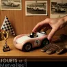 Voiture de course Viglietta Playforever Dottie, jouet design vintage rose pâle avec numéro 9, une main la manipulant, sur une étagère avec gants en cuir et petit trophée.