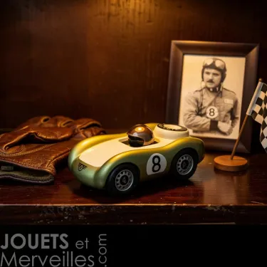 Voiture de course Viglietta Playforever Birdy, design vintage, numéro 8 sur le côté, sur une étagère avec casque, gants et photo d’époque en arrière-plan.
