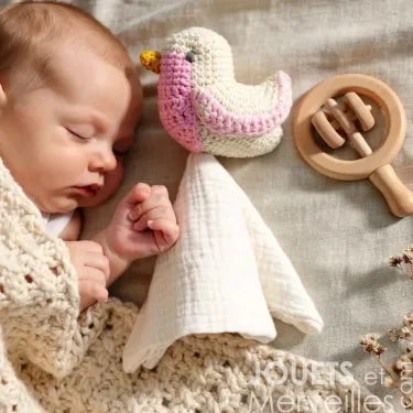 Doudou mésange rose Myum en coton bio près d’un bébé endormi, avec un voile blanc et un hochet en bois à côté.