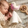 Doudou mésange rose Myum en coton bio près d’un bébé endormi, avec un voile blanc et un hochet en bois à côté.