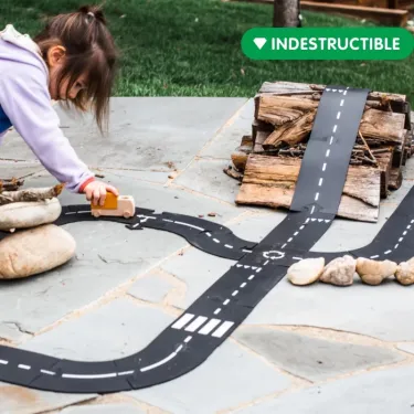 Circuit de route flexible Waytoplay, coffret Autoroute 24 pcs: enfant joue avec son circuit, bandes noires et lignes blanches, autour d’un petit tas de bois.