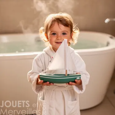 Sloop Tirot | Petit bateau jouet en bois pour enfants: jeune enfant souriant tient un petit voilier en bois dans une salle de bain.