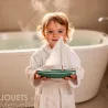 Sloop Tirot | Petit bateau jouet en bois pour enfants: jeune enfant souriant tient un petit voilier en bois dans une salle de bain.