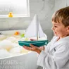 Petit garçon tenant un bateau-jouet en bois Sloop Tirot avec voiles blanches près d’une baignoire.