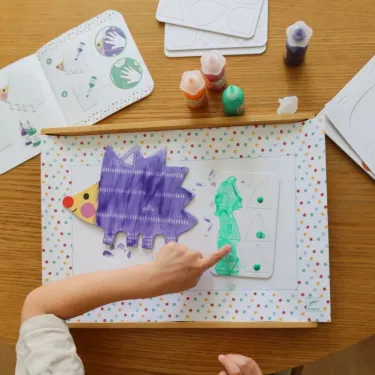Enfant utilisant la peinture à doigts magique Petites formes DJECO 8984 pour colorier une page avec animal en papier coloré sur fond à pois.