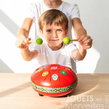 Tambour à languettes DJECO 6039 Metal Drum rouge en jeu sur table, enfant en arrière-plan, mains levées.