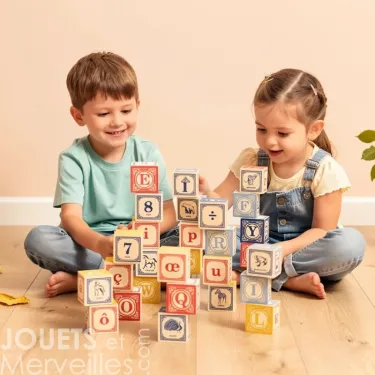 Deux enfants assis par terre jouent avec des cubes alphabet Uncle Goose, formant une pile colorée sur un sol en bois.