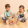 Deux enfants assis par terre jouent avec des cubes alphabet Uncle Goose, formant une pile colorée sur un sol en bois.
