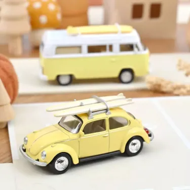 Miniature Volkswagen Coccinelle 1303 1973 NOREV 1/43 en jaune pastel avec planches de surf sur le toit, devant une mini caravane jaune.
