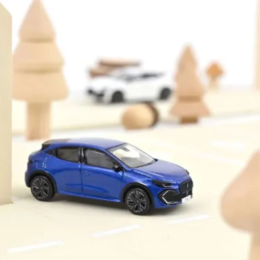 Renault Clio VI 2026 NOREV 1/64 en bleu iron, mini voiture Citadine sur une route légère, décorée de sapins en bois.