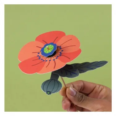 Fleur des champs en papier découpé, rose avec bouton central bleu, tenue par un bâtonnet tenu par une main contre fond vert. Produit: kit créatif Pirouette Cacahouète.