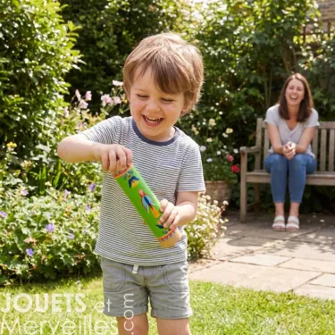 Enfant souriant faisant t/interagir un bâton de pluie en bois vert, jardin en arrière-plan et une femme assise sur un banc au fond.