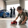 enfant jouant au sol dans un garage avec sa voiture Baghera, réplique de Porsche 911. Véritable Porsche 911 bleue en arrière plan.