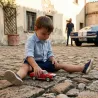 Petit garçon en chemise bleu joue avec une voiture Baghera rouge sur une rue pavée, avec une voiture Mustang classique en arrière-plan.