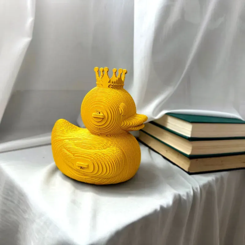 Canard jaune en forme de luciole avec couronne, sculpture en spirales sur fond blanc, posé sur une nappe près de livres.