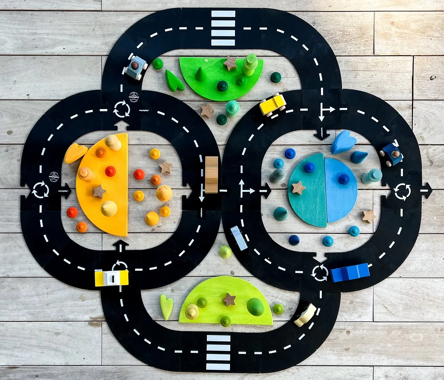 Circuit de voiture en bois géant sur parquet, traceré en boucle noire avec routes blanches et intersections, éléments colorés et pièces miniatures placés dessus.