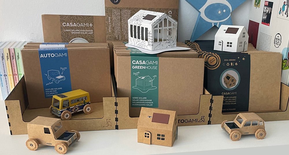 Collection de petites maquettes en carton et bois, emballages marque Casagam, camions et maisons miniatures sur fond blanc.