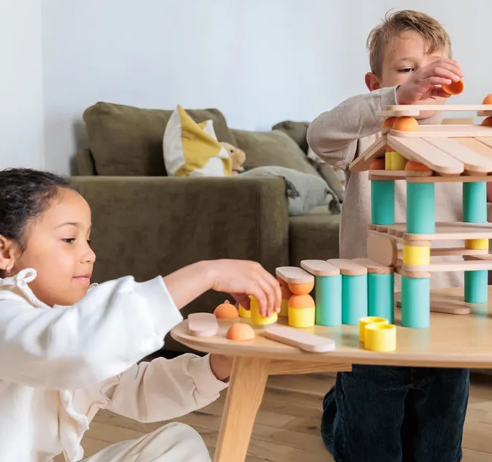 Deux enfants jouent avec des pièces de construction en bois sur une table, créant une tour colorée et des éléments empilables en bois naturel et silicone de couleur.
