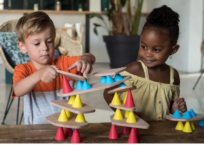 Deux enfants jouent avec des pierres et cônes colorés sur des plateaux en bois, empilant une structure en équilibre.