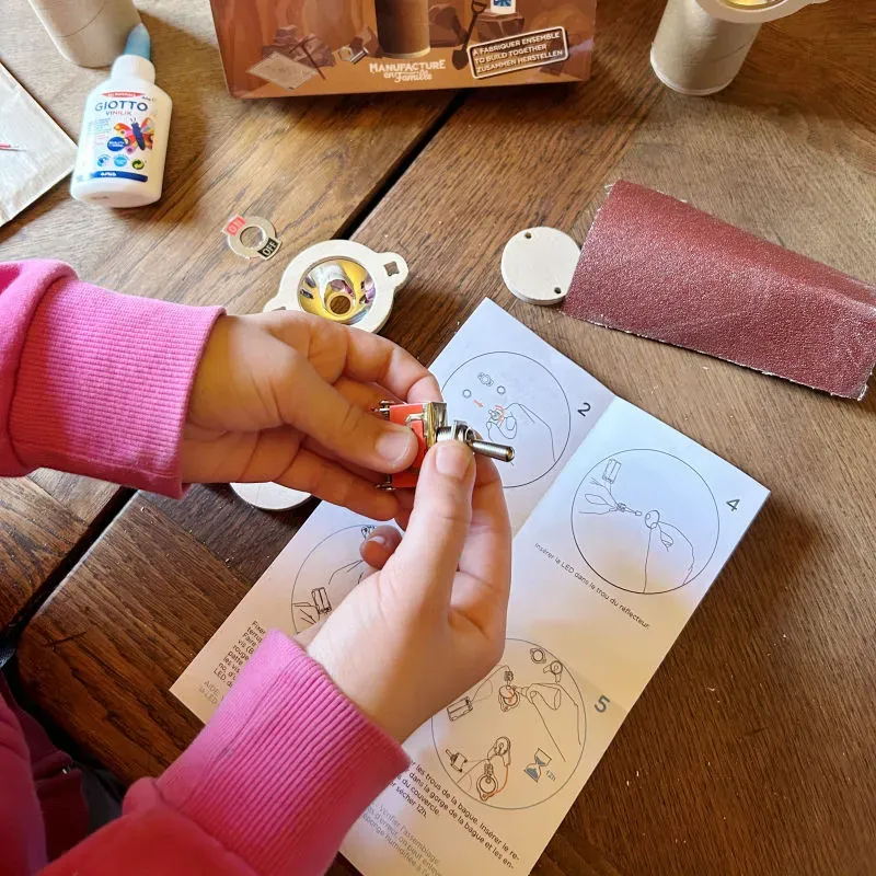 Enfant assemble des pièces miniatures sur une feuille d’instructions pour une activité créative, fabrication d'une lampe. Matériel de bricolage et petit outil métallique sur table en bois.
