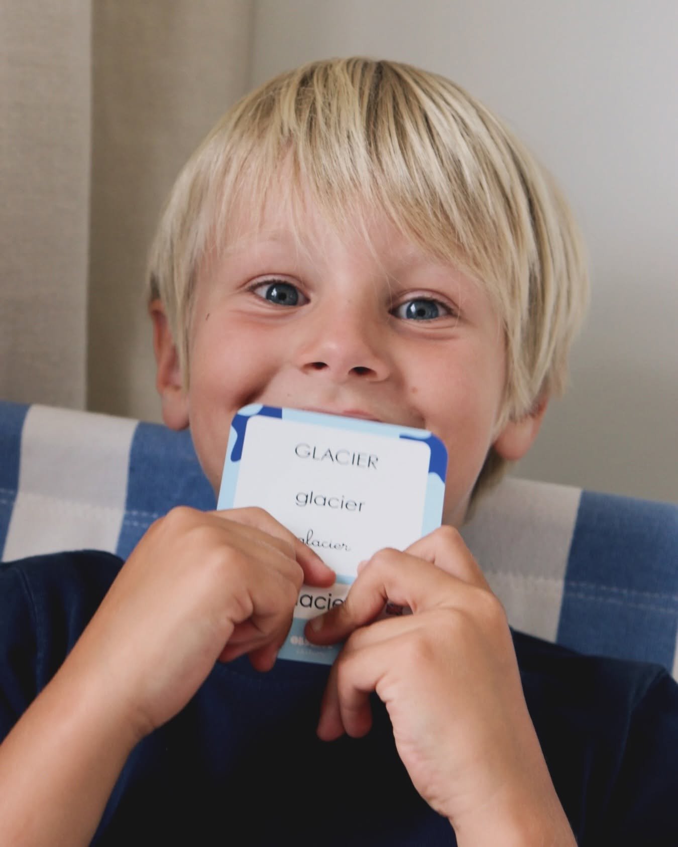Enfant blond souriant tenant une carte bleue en papier près de la bouche, regard souriant et yeux bleus, assis sur un canapé à motifs bleus.