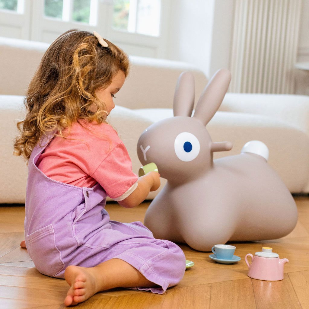 Petite fille assise sur le sol en bois, jouant avec un grand doudou lapin gris et des jouets de thé colorés dans un salon lumineux.
