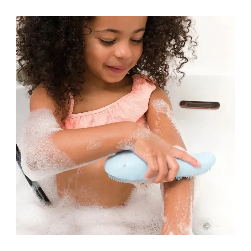 Petite fille se lavant les bras avec une brosse-outil bleue dans la baignoire remplie de mousse et de savon.