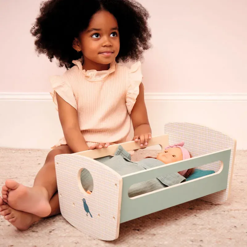 Poupée bébé Poméa Djeco dans un berceau en bois, enfant assise près d’elle, décor pastel. Poupée bébé Poméa Djeco dans un berceau en bois, enfant assise près d’elle, décor pastel.