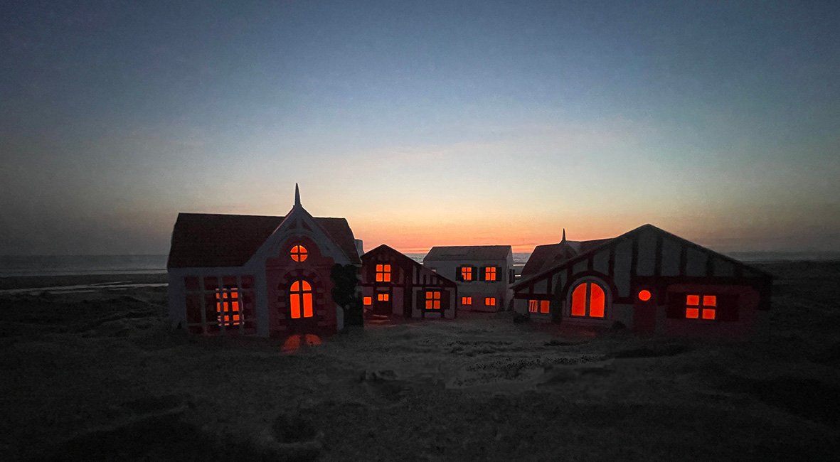 Ville miniature illuminée à la tombée de la nuit sur la plage, maisons éclairées en rouge/orange.