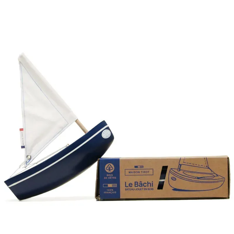 Voilier jouet en bois bleu foncé avec voile blanche et mât, accompagné d’une boîte en carton beige illustrée “Le Bâchi” pour jouets nautiques.